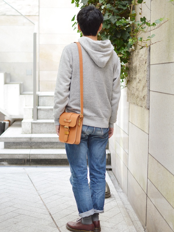 淺棕色皮革製成的單肩包，配有金色皮扣和可調式肩帶，由男性斜背於灰色連帽衫和牛仔褲外。