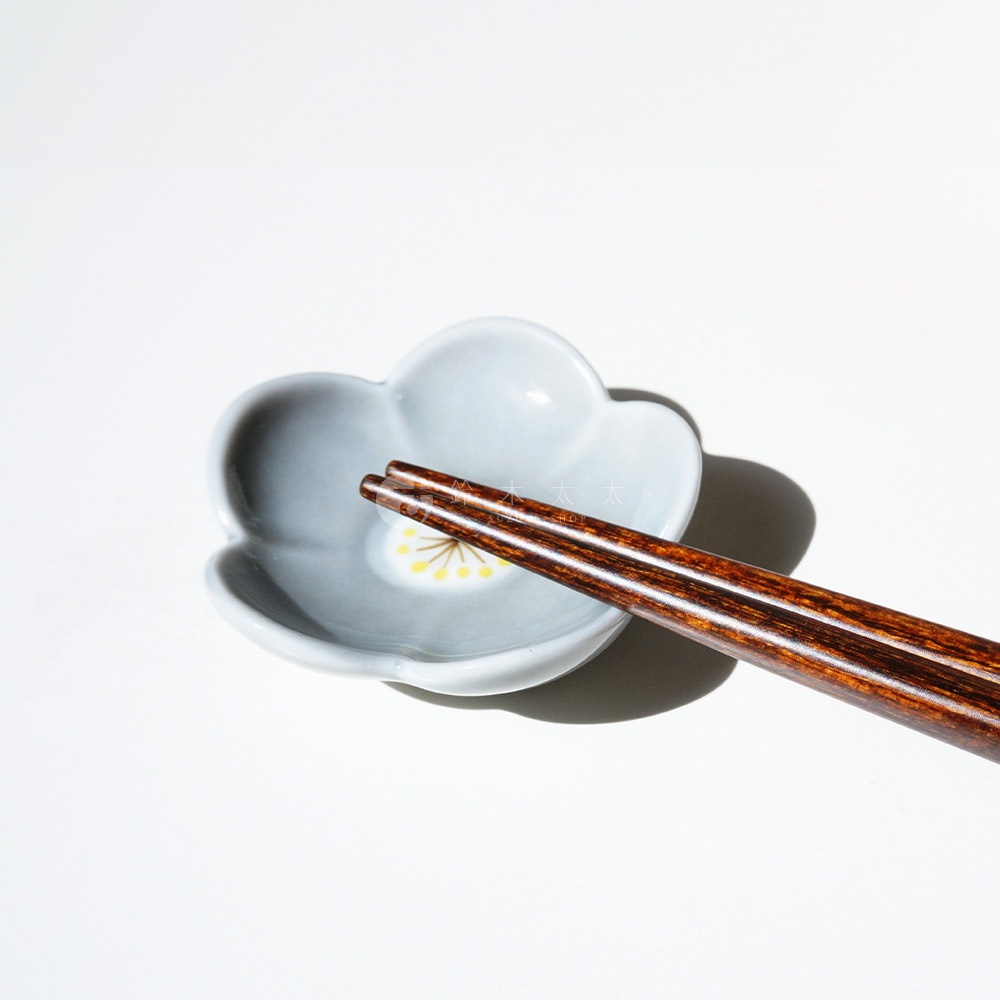 一雙深棕色木質筷子，放在一個淺灰色、花朵造型的陶瓷筷架上，筷架中心有黃色花蕊圖案。