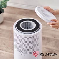 白色圓筒形空氣清淨機，頂部可見濾網，旁邊有一隻手正拿起白色圓形頂蓋。機身有細密網孔，底部印有IRIS OHYAMA標誌。