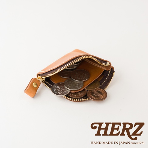 一個職人手作的日本HERZ淺棕色皮革零錢包，採拉鍊開口設計，包內裝有數枚不同面額的日本硬幣，整體造型簡約精巧，右下角印有品牌標誌與產地資訊。