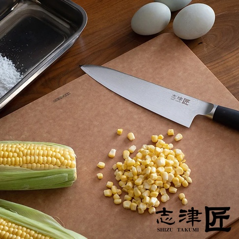 銀色不銹鋼萬用刀，黑色刀柄，刀身上有細緻紋路，置於咖啡色砧板上，旁邊散落著玉米粒，左側有一未剝殼玉米，上方有不銹鋼盤及兩顆白色雞蛋。