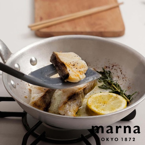 marna TOKYO 1872，一塊煎好的魚肉被灰色矽膠刮刀從鍋中取出，鍋裡還有其他魚肉、迷迭香和檸檬片。