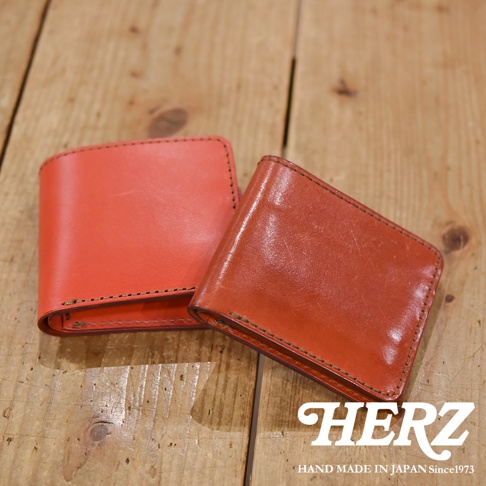 兩款皮革二折短夾，一款為亮橘色，另一款為較深的橘棕色。兩者皆為橫式設計，以手工縫線包邊，材質為皮革。