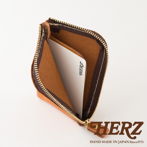 一個日本HERZ職人手作棕色皮革迷你零錢包，以俯視角度呈現半開狀態，內部設有皮革分隔層並放置了一張印有HERZ標誌的卡片，拉鍊處呈現L型設計，右下角印有HERZ品牌名稱與相關標語。