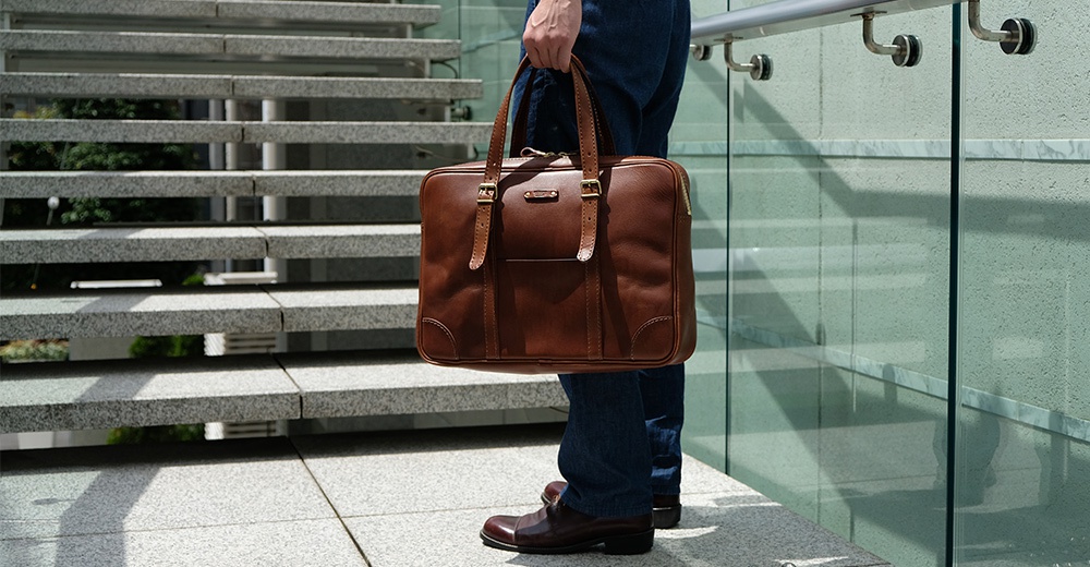 棕色皮革製成的職人手作公事包，可肩背或手提。包款有雙提把、拉鍊開合，正面有磁釦口袋與金屬扣帶裝飾。