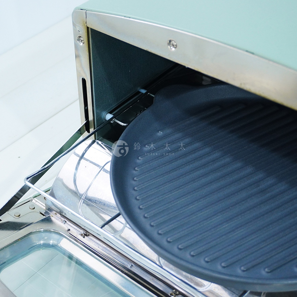 淺綠色烤箱內，放置著一個黑色、圓形、帶有波浪紋路的陶製烤盤，烤盤置於不鏽鋼烤架上，烤架下方是烤箱的透明玻璃門。