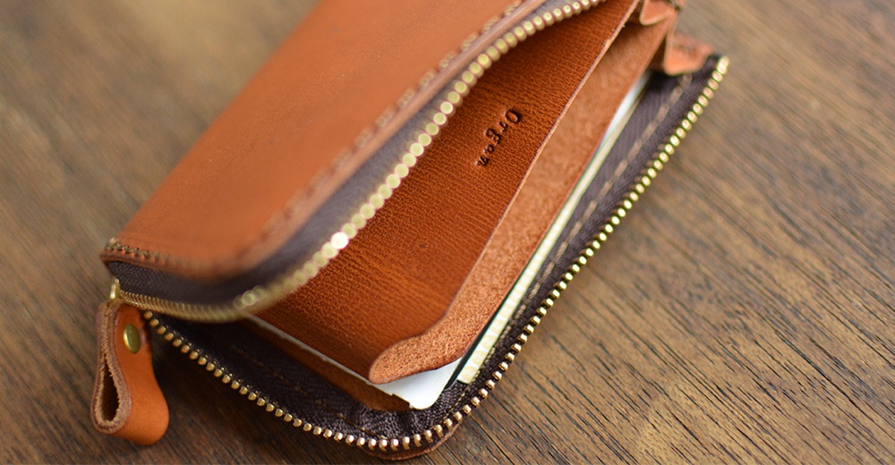 棕色皮革拉鍊短夾，內有卡槽，夾層可見「Organ」燙金字樣，搭配金色拉鍊與黃銅鉚釘，置於木紋桌面上。