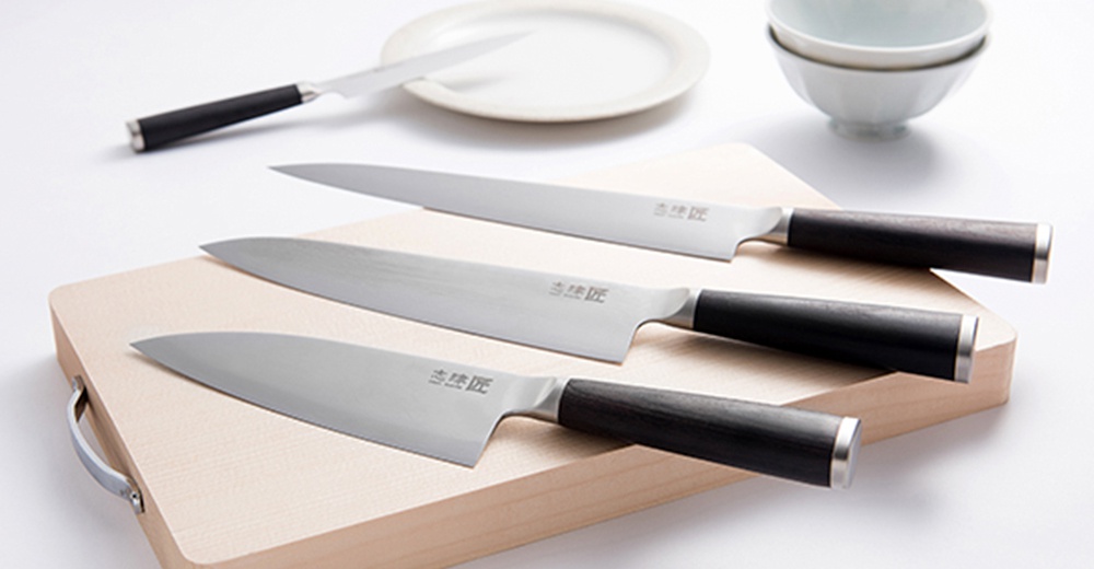 三把銀色金屬刀刃、黑色木柄的水果刀，其中兩把橫放於淺色木質砧板上，另一把斜放於後方，背景有白色盤子和碗。