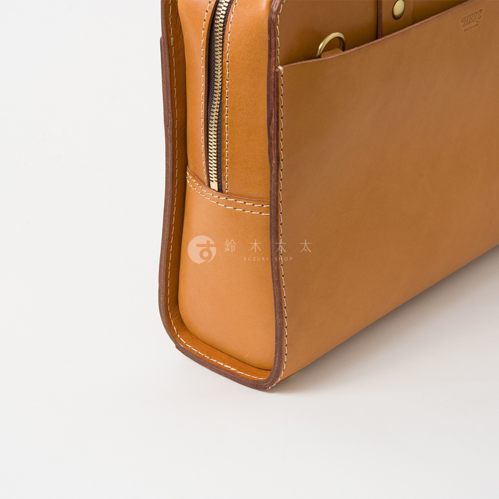 職人手作皮革雙口袋兩用商務公事包，以淺棕色皮革製成，擁有金色拉鍊與皮革縫線細節，包體側面呈現圓潤的皮革邊緣。