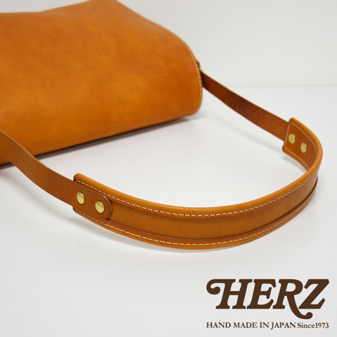 皮革復古翻蓋郵差包，材質為天然皮革，顏色為駝色，包包提把部分特寫，縫線清晰，金屬鉚釘裝飾。