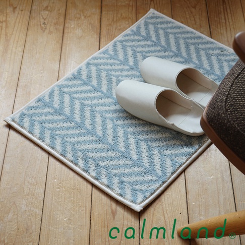 淺藍色與白色相間的羽毛圖案玄關地墊，搭配一雙乳白色室內拖鞋，擺放在淺木紋地板上。