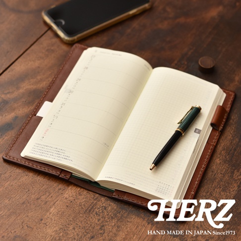 職人手作皮革旅行記事本書套，焦糖棕色，內含一本打開的日誌，一本綠色封面的筆記本，一支綠色與金色筆夾的鋼筆，以及一部黑色手機。