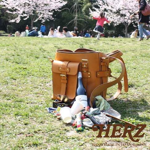 職人手作皮革大型三用旅行後背包，天然棕色皮革，帶有兩個前翻蓋口袋、金屬扣環和側邊揹帶。旁邊擺放著酒瓶、塑膠杯和野餐用品。