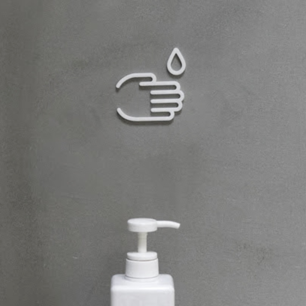 白色洗手標示牌，圖案為一隻張開的手掌上方有一滴水，下方搭配白色按壓式皂液器，整體設置於灰色水泥牆面上。