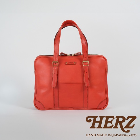 鮮紅色皮革製成的手提肩背包，雙提把，附有可調節長度的肩帶扣環，包身正面有一個大口袋，底部有加固皮革，搭配金色五金配件。