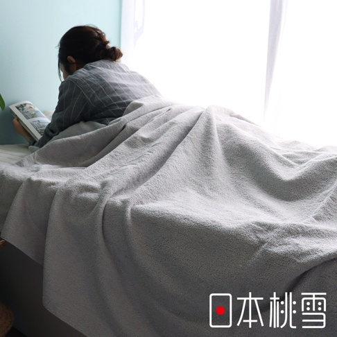 淺灰色雙面毛圈超長棉毛巾被，材質柔軟，覆蓋在一位正在閱讀的女性身上，呈現居家休閒氛圍。