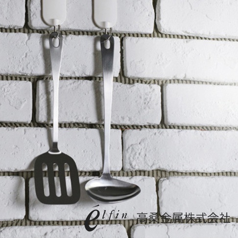 一組日製不鏽鋼廚具，包含一個帶有長柄和圓形湯勺的湯杓，以及一個帶有長柄和方形濾網的鍋鏟，兩者皆掛在白色磚牆上。