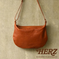 一個棕褐色皮革製成的單肩半月包，包身柔軟，搭配皮革肩帶，懸掛在淺褐色的背景前。