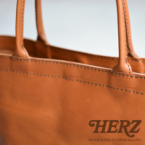 HERZ 品牌字樣和 HAND MADE IN JAPAN Since1973 字樣出現在一個淺棕色皮革托特包的近景特寫中，包包表面有精緻的縫線細節。