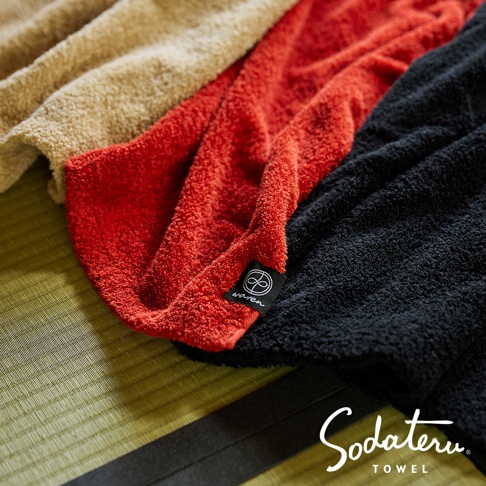 三條不同顏色的毛巾，分別是卡其色、紅色和黑色，疊放在綠色的藺草墊上，紅色毛巾上有一個寫著 wa/en 的小標籤