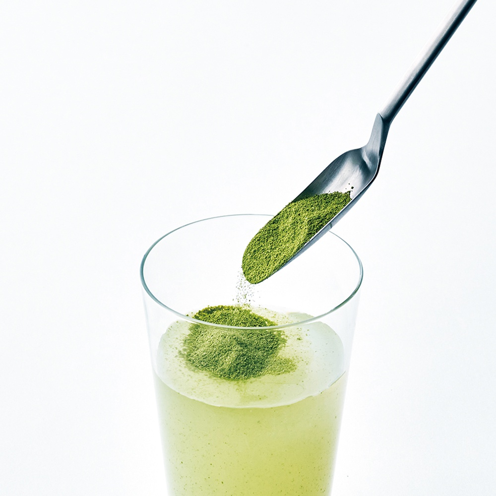 日製金屬長柄細勺，盛著綠色粉末，正從透明玻璃杯上方傾倒。杯中是淺綠色液體，部分粉末已落在液體表面。