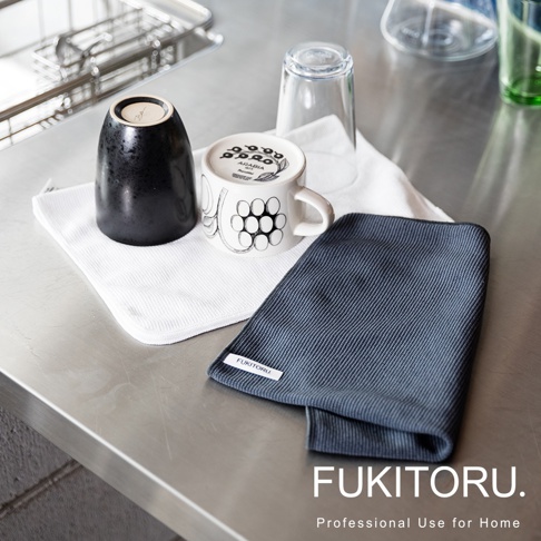 兩條超極細纖維擦拭布，一條為白色，一條為深藍色，表面有細緻的織紋。圖片中還有一黑一白兩個杯子，以及兩個玻璃杯，皆放置在金屬檯面上。