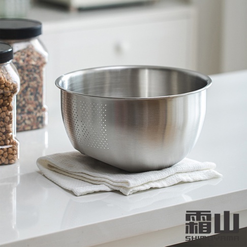 一個斜底不鏽鋼瀝水料理盆，盆壁有密集的小孔，放置在白色毛巾上，背景是廚房檯面和裝有豆子的儲物罐。