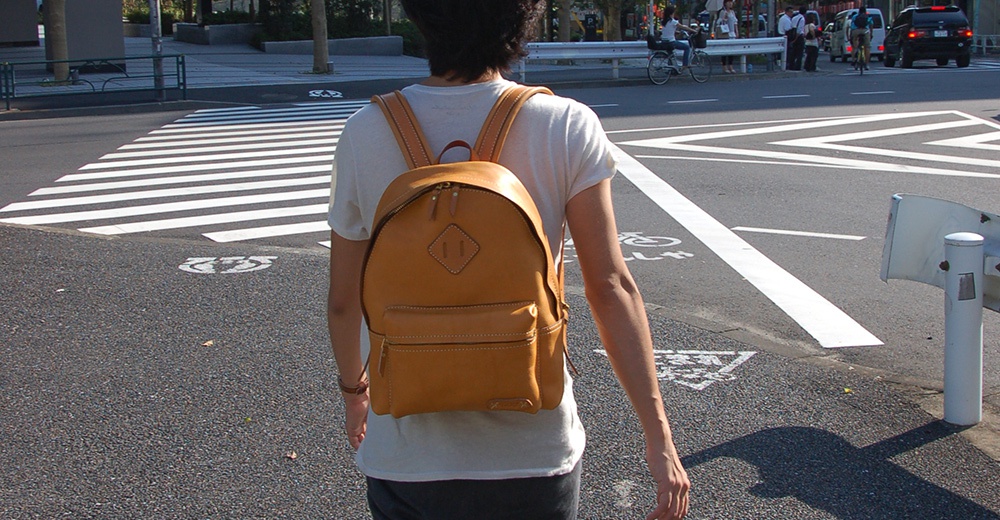 一款棕褐色皮革後背包，正面有一個方形裝飾和一個拉鍊口袋，配有雙肩帶，由一人背負於街頭。