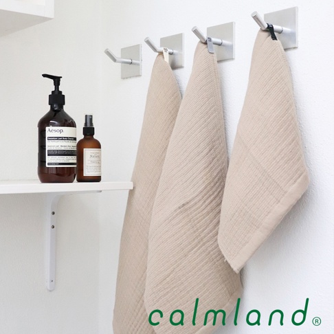 三條淺米色無撚紗質夾心長毛巾，質地柔軟，懸掛於白色牆面上的銀色掛鉤。左側架子上擺放著兩瓶棕色瓶身的清潔用品。