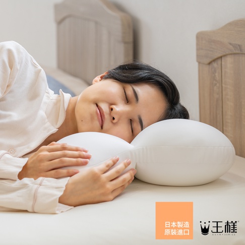 一位女性穿著米白色睡衣側臥在床上，頭部舒適地枕在一個設計獨特的白色王樣夢枕上，雙手輕輕交疊環抱枕頭邊緣，畫面右下角呈現王樣品牌標誌與日本製造原裝進口的文字資訊。