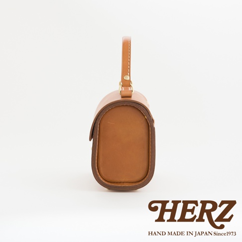 職人手作皮革復古杜勒斯手提包，展示其側面，包身呈圓潤的橢圓形，採用天然棕色皮革製成，配有單一皮革提把及金屬扣環，整體風格簡約復古。