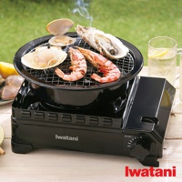 黑色雙重防風卡式爐，上方有烤網，網上烤著鮮蝦、扇貝和生蠔，冒著微微煙霧，爐身印有 Iwatani 商標。
