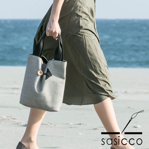 一位女性提著一個直式托特包，包身為黑白細條紋的刺子織紋理，配有黑色提把和一個木質圓形鈕扣，整體風格簡潔。