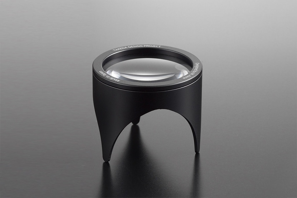 黑色鋁製閱讀放大鏡，2.5倍放大，鏡筒周圍刻有「TAKEDA DESIGN PROJECT」、「Designed by M. Akira」及「Made in Japan」字樣。