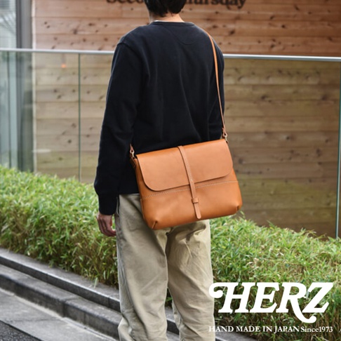 一名男子身揹棕色皮革馬鞍包，走在戶外，包身上有 HERZ 品牌標誌。