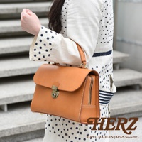 HERZ 職人手作淺棕色皮革多功能收納單肩包，採用經典鎖頭翻蓋設計，側邊有拉鍊收納，並印有品牌 logo 及日文標語 HAND MADE IN JAPAN Since1973。