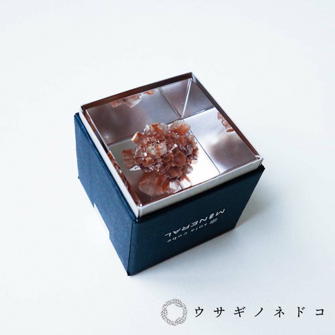 深藍色方形外盒內，置放著一個閃亮的金屬盒，盒內有一簇紅褐色、多稜角的霰石礦石，礦石表面呈現結晶狀。