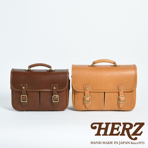 兩個職人手作皮革單肩包，一個為深棕色，另一個為淺棕色。兩款包均為翻蓋設計，配有兩個皮帶扣和金屬插扣，頂部設有提把。