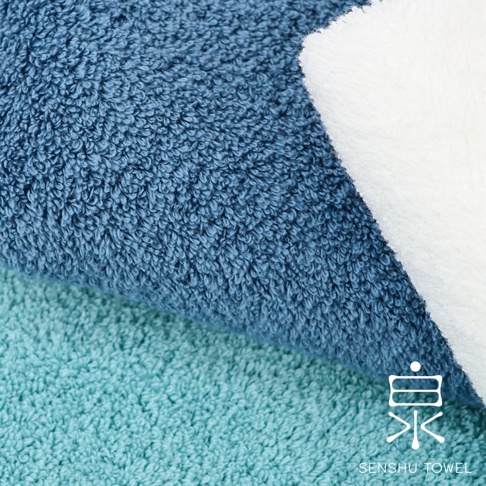 特寫鏡頭展示了兩條毛巾的材質，一條是藍色，另一條是淺綠色，兩條毛巾的絨毛都清晰可見。右側疊放著一條白色毛巾，其絨毛質地略有不同。右下角有白色「泉」字標誌和「SENSHU TOWEL」字樣。