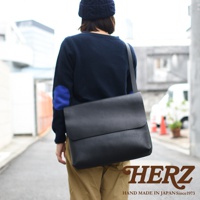 黑色皮革經典郵差包，配有可調節肩帶，展示於穿著海軍藍毛衣和卡其色褲子的模特身上。