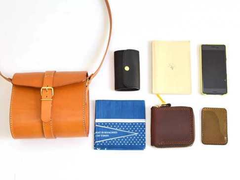 職人手作皮革皮扣單肩包，採用鞣製皮革製成，呈現自然棕色調，搭配金色皮帶扣和縫線。包款為圓筒狀，配有可調式長背帶。周圍擺放有黑色皮夾、黃色筆記本、手機、藍色印花手帕、咖啡色拉鍊皮夾和卡夾。