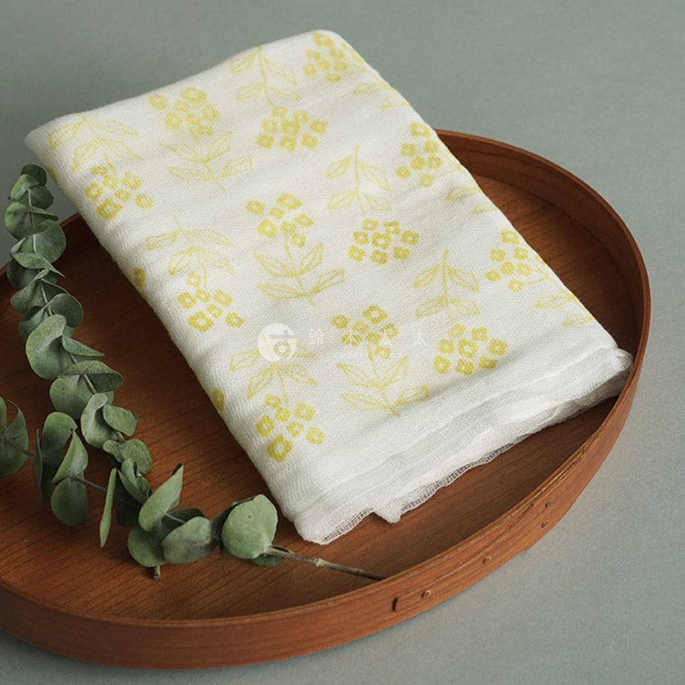 一條白色純棉花草紗布大拭巾，印有黃色小花圖案，置於木質圓盤中，旁有一枝綠色植物。