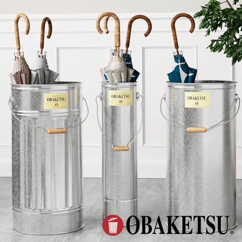 OBAKETSU 手工製傘桶，左邊較寬的傘桶內有條紋和灰色傘，中間和右邊較窄的傘桶內有花紋傘，傘桶均為銀色金屬材質，附有木製提把和標籤。