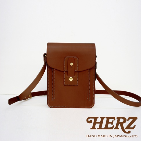 棕色皮革製成的直立式斜背包，正面有兩個金屬釦和縫線裝飾，附有可調整的長背帶。