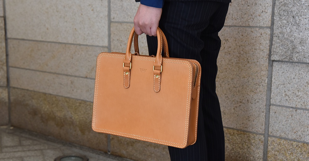 淺棕色皮革手提商務公事包，配有金色金屬扣環和拉鍊，以及對比色縫線。
