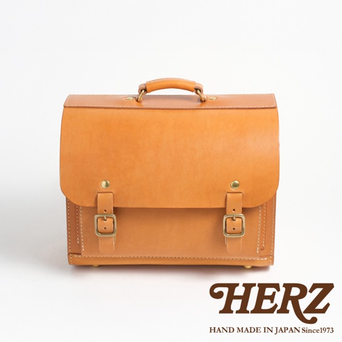 職人手作皮革橫式書包，帶有兩個金屬釦環和提把，品牌HERZ標誌在右下角