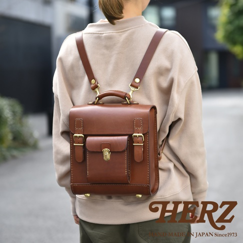 職人手作的經典學院風迷你後背包，採用光滑的棕色皮革製成，配有黃銅色金屬扣環和旋轉鎖。背包設計方正，前方有一個帶有翻蓋的小口袋，頂部有一個手提把和可調節的肩帶。