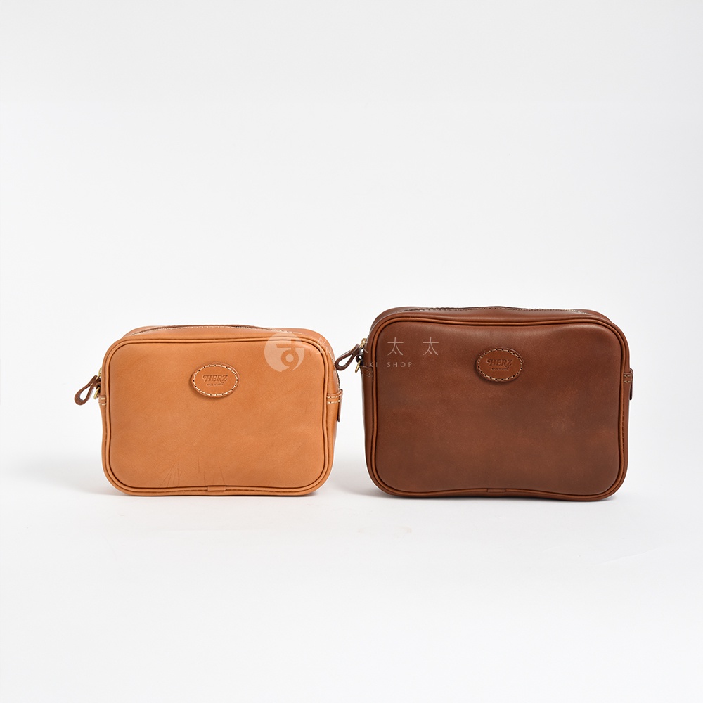 職人手作皮革可拆式扣環萬用手提包，左為淺棕色，右為深棕色，兩款皆為拉鍊開合，正面有品牌圓形皮革標誌。