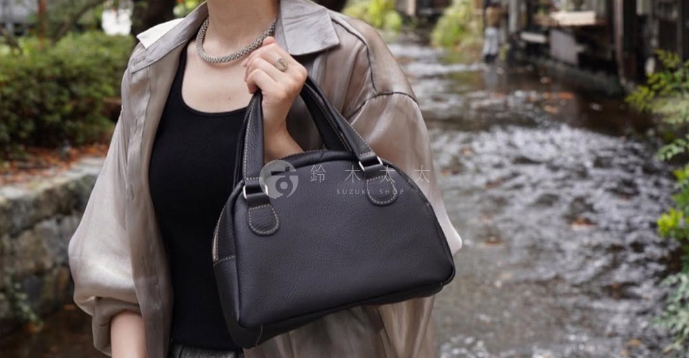 深棕色皮革迷你手提包，半月形設計，配有雙提把和拉鍊開合。包身有縫線細節，展現職人手工質感。