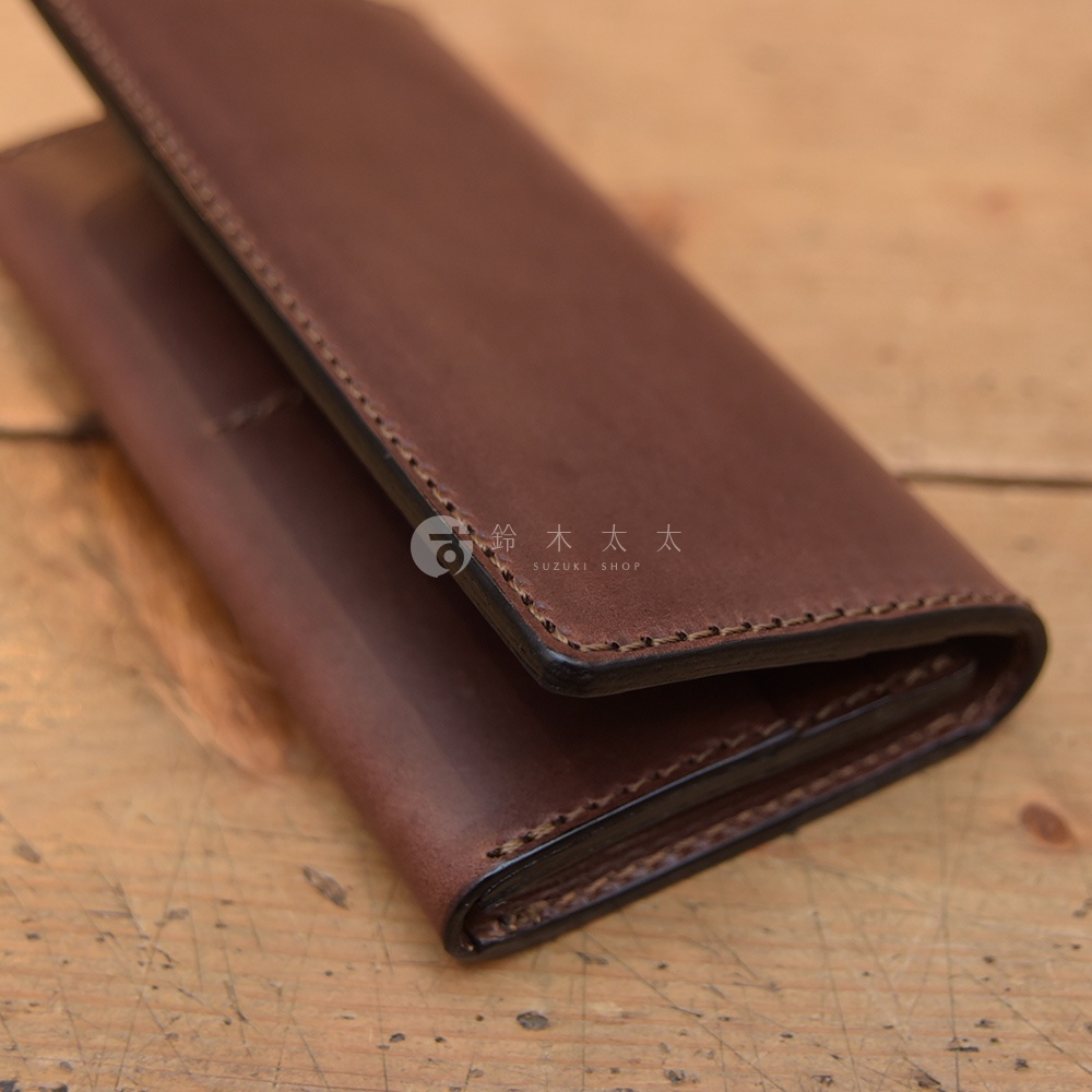 這款日本HERZ手工製作的Latigo軟皮革長夾，呈現深褐色的溫潤質感，以細緻的手工縫線裝飾邊緣，表面印有鈴木太太標誌，特寫視角展示了皮革折疊後的厚實層次與精湛工藝。